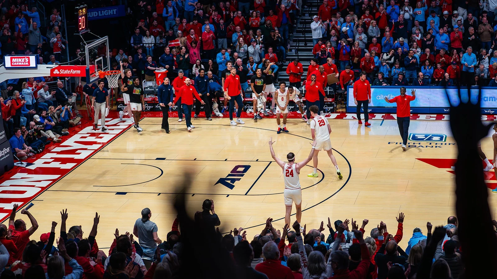 A wide-angle view of a packed basketball arena captures the home crowd celebrating as two players in white uniforms stand on the court with their arms raised toward the fans. Teammates and coaches along the bench leap to their feet in celebration, while spectators throughout the arena cheer and applaud, creating a high-energy atmosphere during the game.