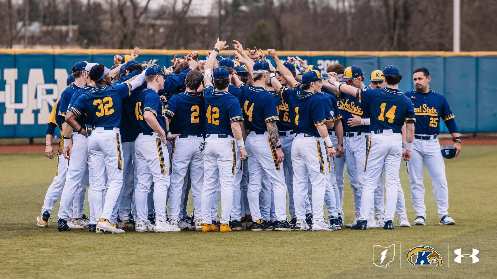 **The Kent State Golden Flashes baseball team gathers in a pregame huddle on the outfield grass at home, with players in navy and gold Under Armour uniforms raising their hands and fingers together in a unified team cheer. Visible jersey numbers include 32, 2, 42, 44, 14, and 25, with "Kent State" lettering across the backs of several players. A coach stands to the right holding a helmet as the group breaks together, with the outfield wall and scoreboard visible in the overcast background.**