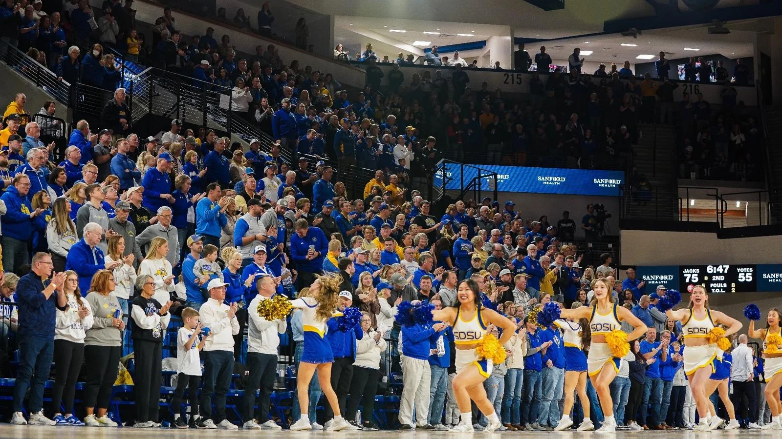 Cheerleaders in white uniforms on the court while fans in the stands in yellow and blue attire applaud