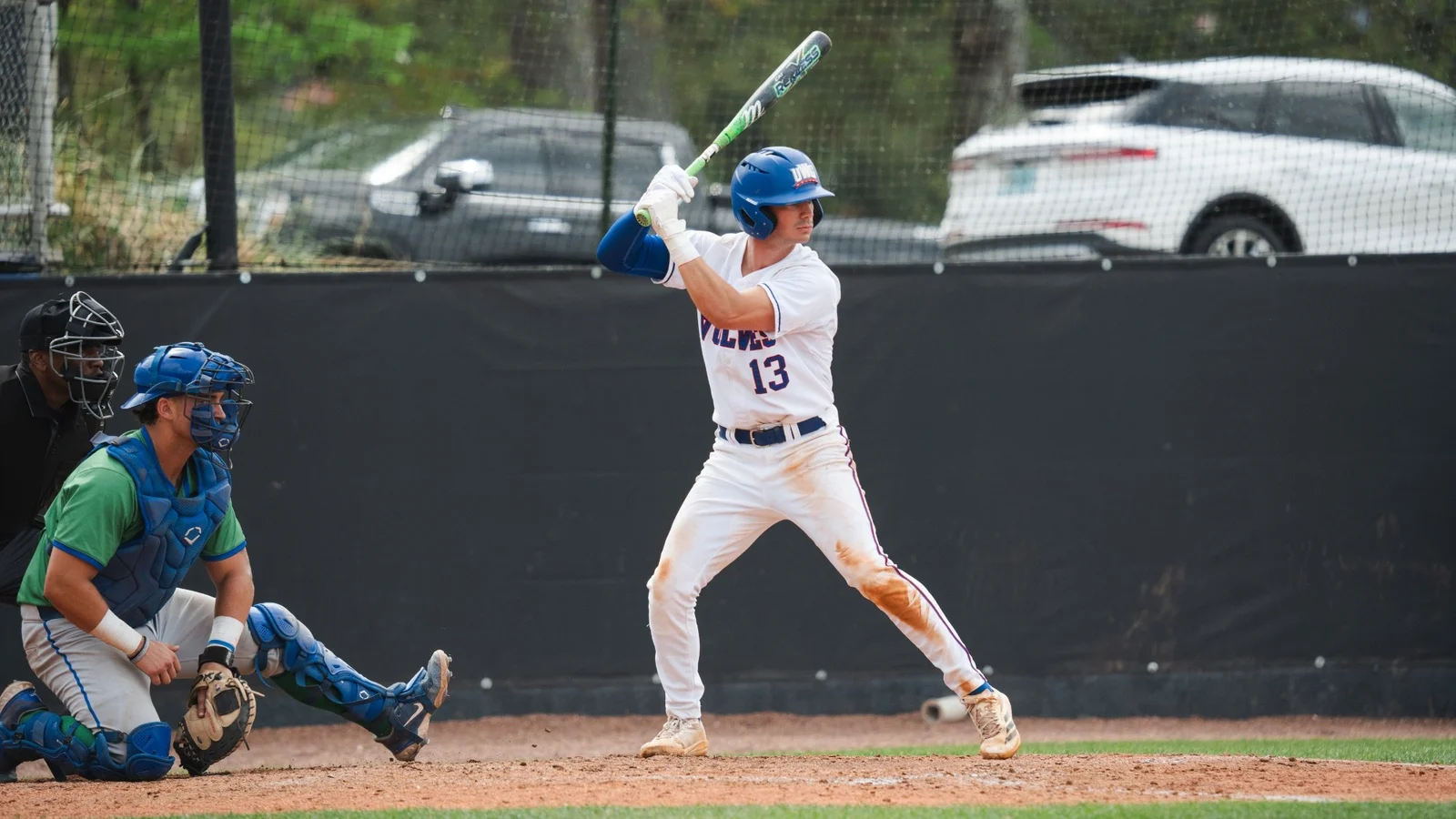 Baseball player in green jersey and blue gear kneeled down waiting to catch pitch. Player in white uniform with blue helmet next to him waiting to swing bat